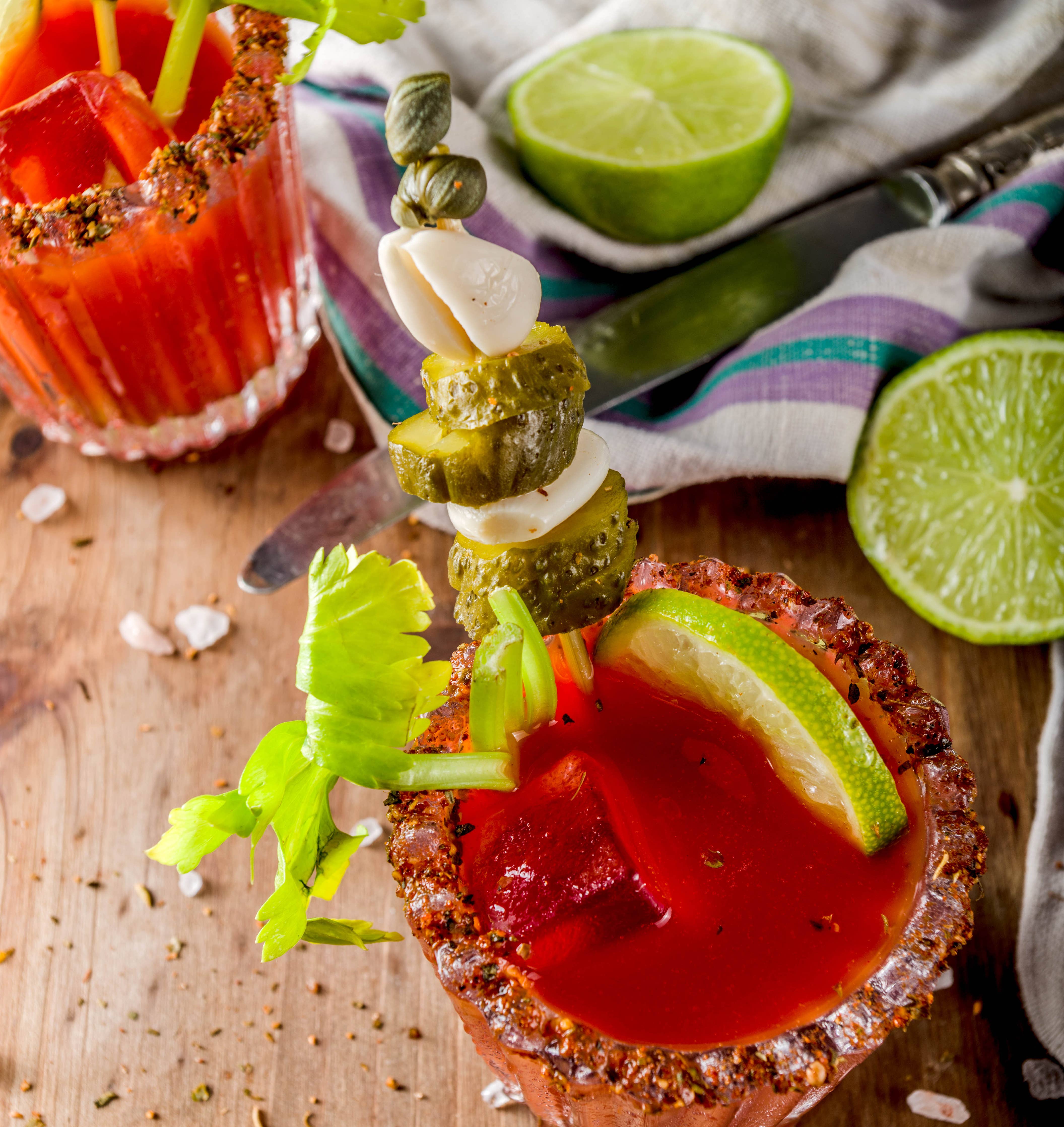 Bloody Mary cocktail with garnish on a wooden surface with limes and a striped towel.