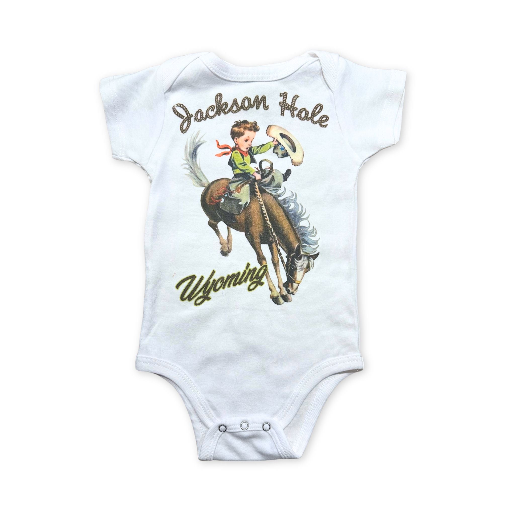 White baby onesie with little cowboy riding a bucking bronco. Text in a rope design reads " Jackson hole wyoming"