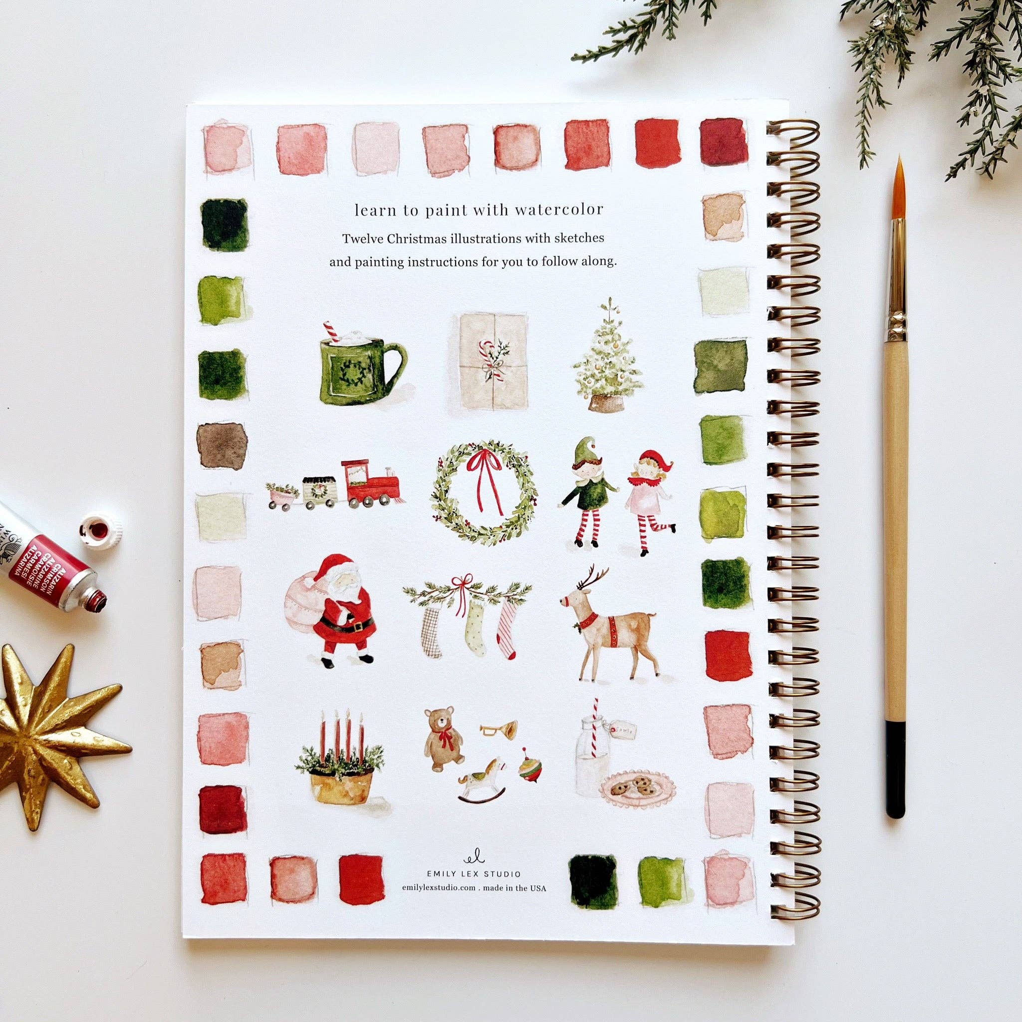 Artistic watercolor illustrations of Christmas-themed designs on a spiral-bound book with a paintbrush and watercolor bottles.