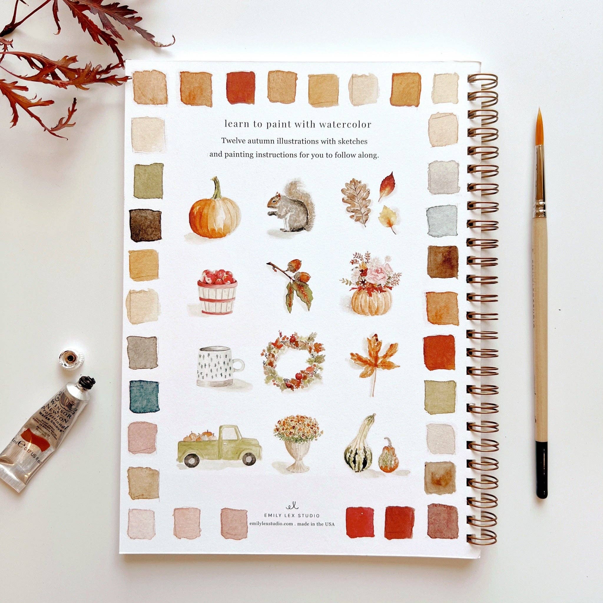 Artbook with watercolor illustrations of autumn elements on a white surface with a paintbrush and water bottle.