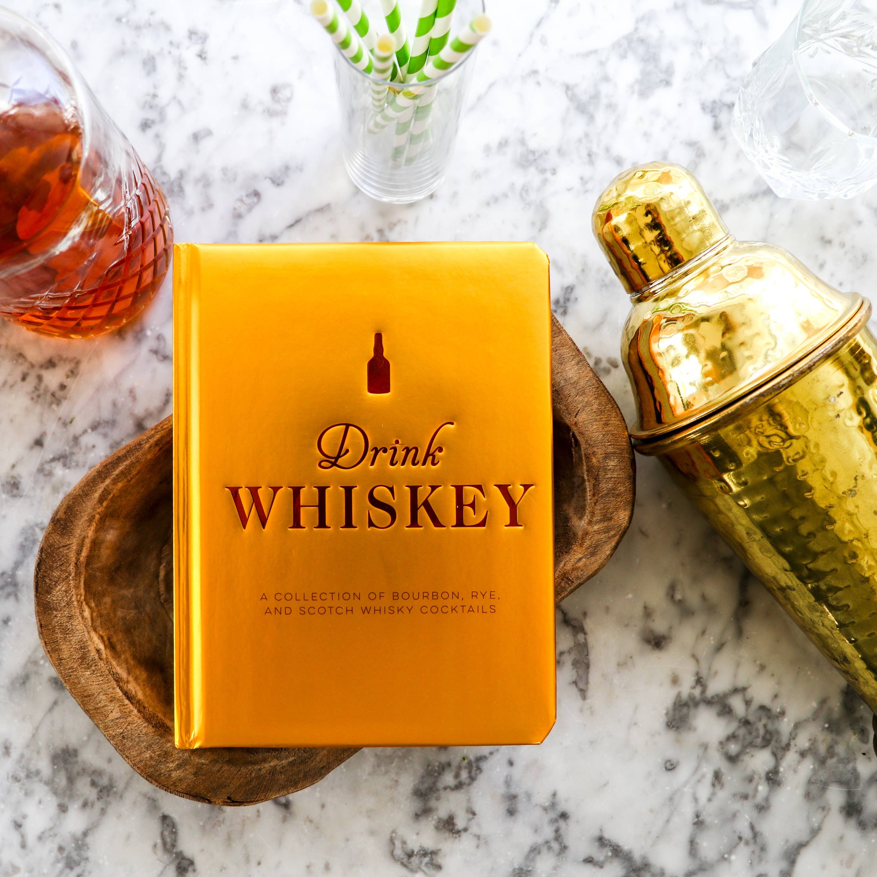 Yellow book titled 'Drink Whiskey' with a glass of whiskey and gold shaker on a marble surface