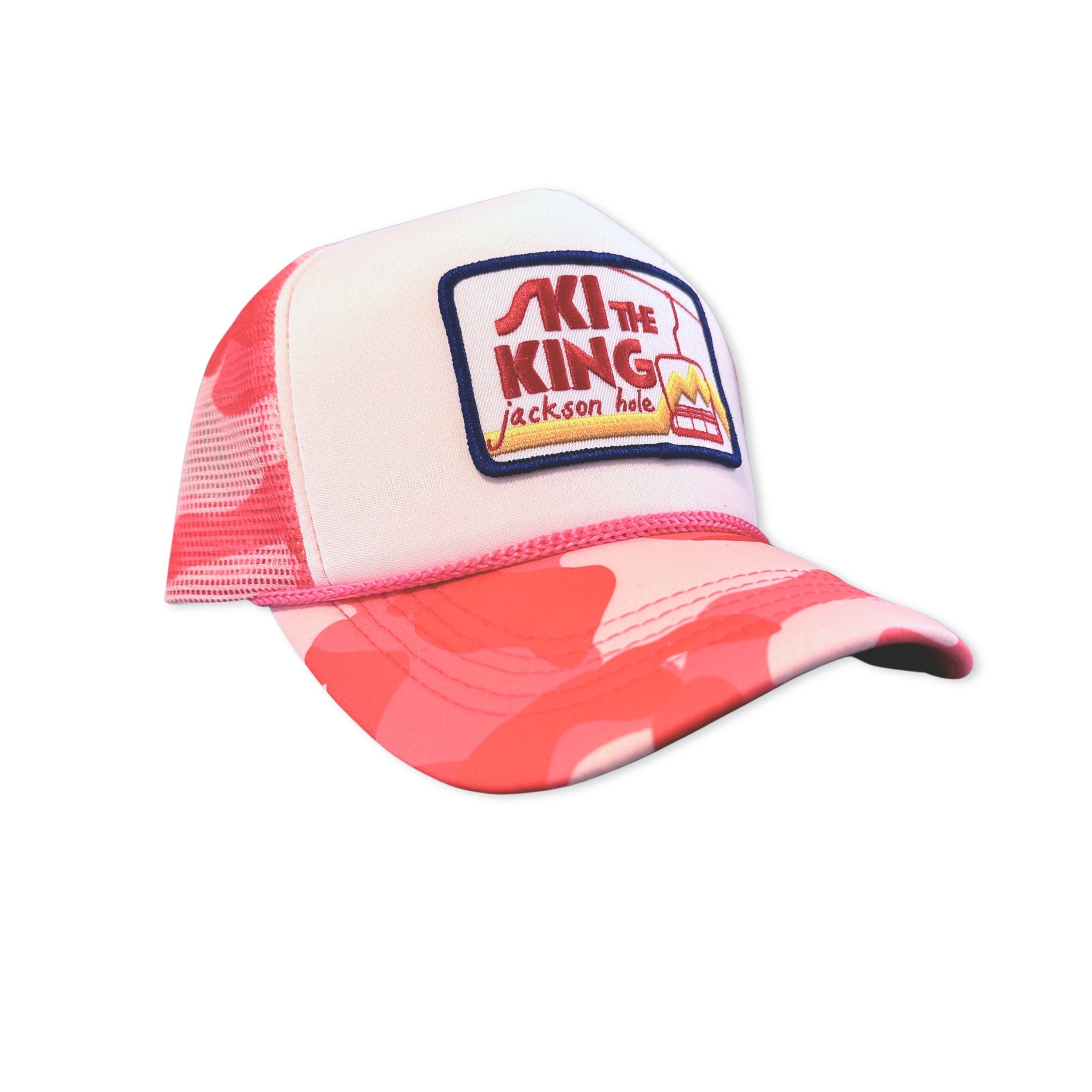 A pink camo foam trucker hat that has a patch with a chairlift ad mountains. Text reading "Ski the King Jackson Hole"