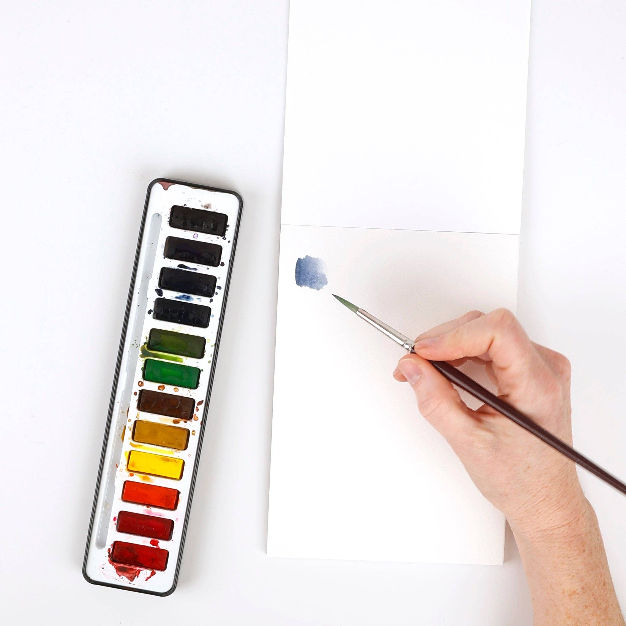 Watercolor paint set with a brush applying paint to a white surface