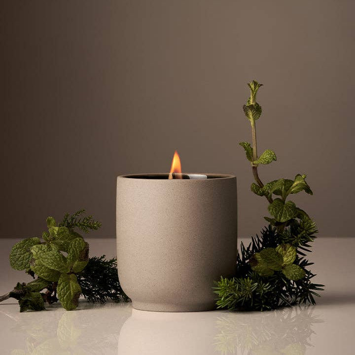 Ceramic candle holder with a lit candle surrounded by greenery on a neutral background