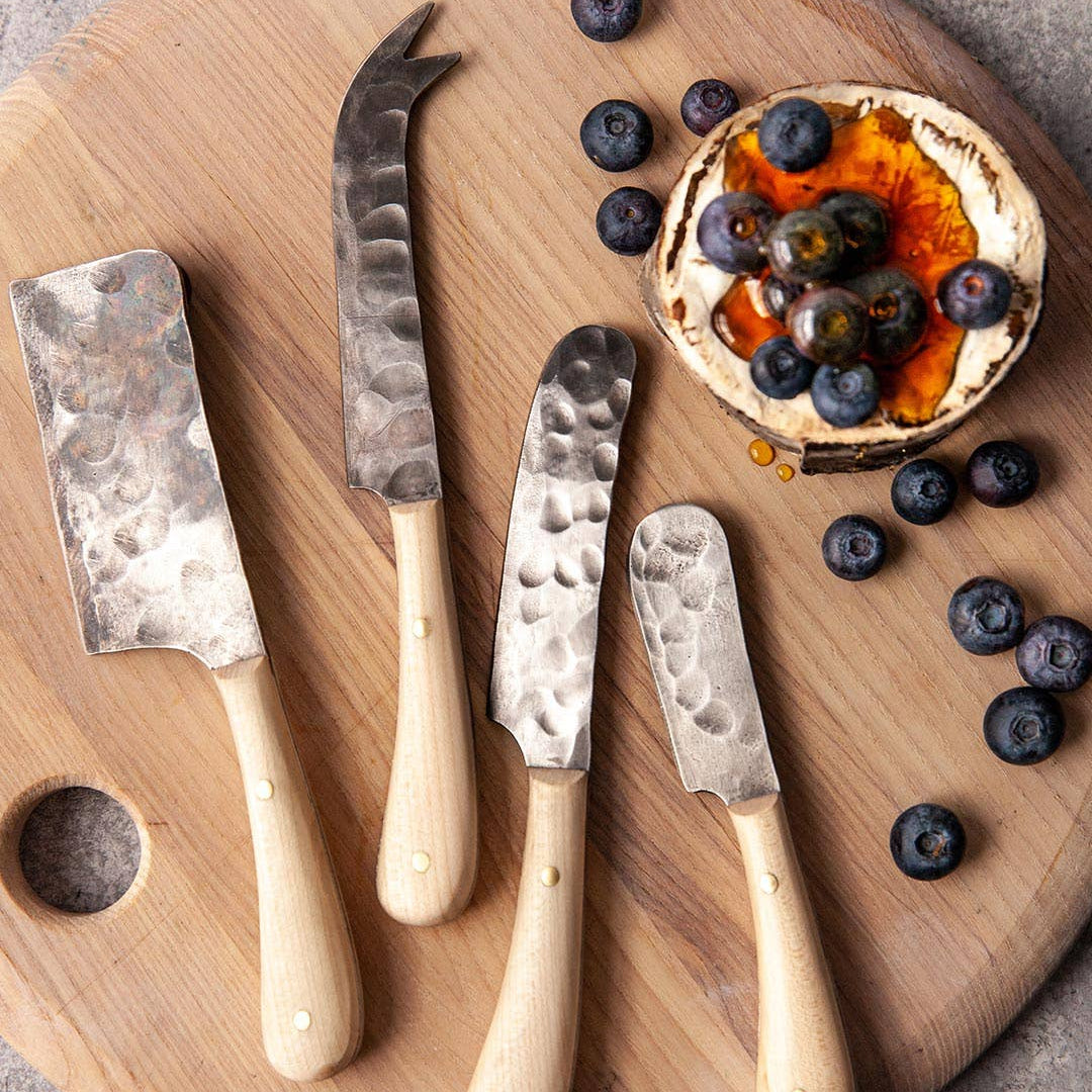 Set of four cheese knives on a wooden board with a small appetizer of blueberries and a round bread slice.