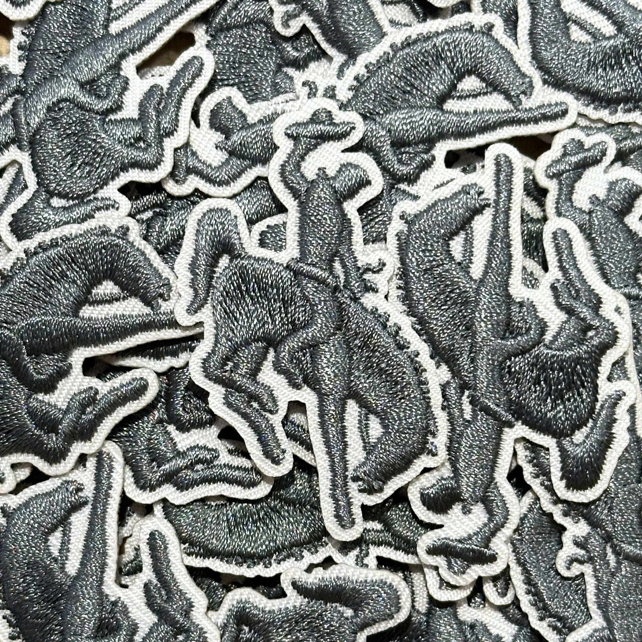 Embroidered patch, showing the silhouette of a bucking bronco and cowboy in grey stitching and white boarder.
