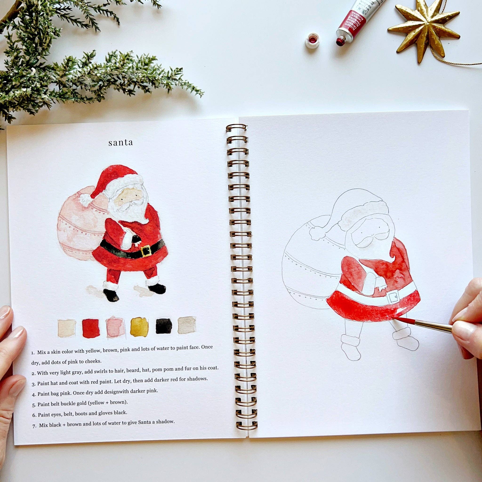 Open book with a drawing of Santa Claus and painting guide on a white surface with Christmas decorations.