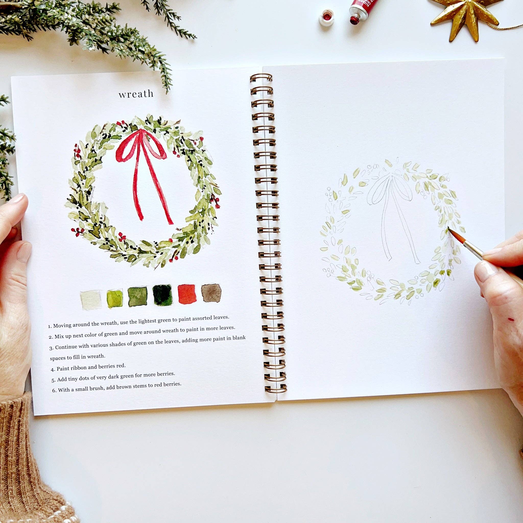 Open book with Christmas wreath illustration and color swatches on a white surface.