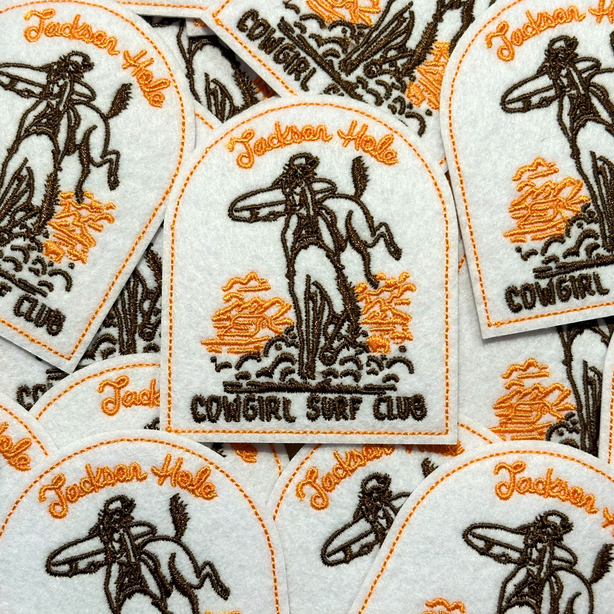 Arched shape patch, showing cowgirl surfer, and cowgirl surf club in brown coloring. Along with Mormon Row. and Jackson Hole in orange coloring.
