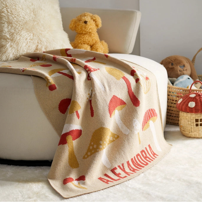 Blanket with mushroom pattern draped over a chair, with a teddy bear and wicker basket in the background.