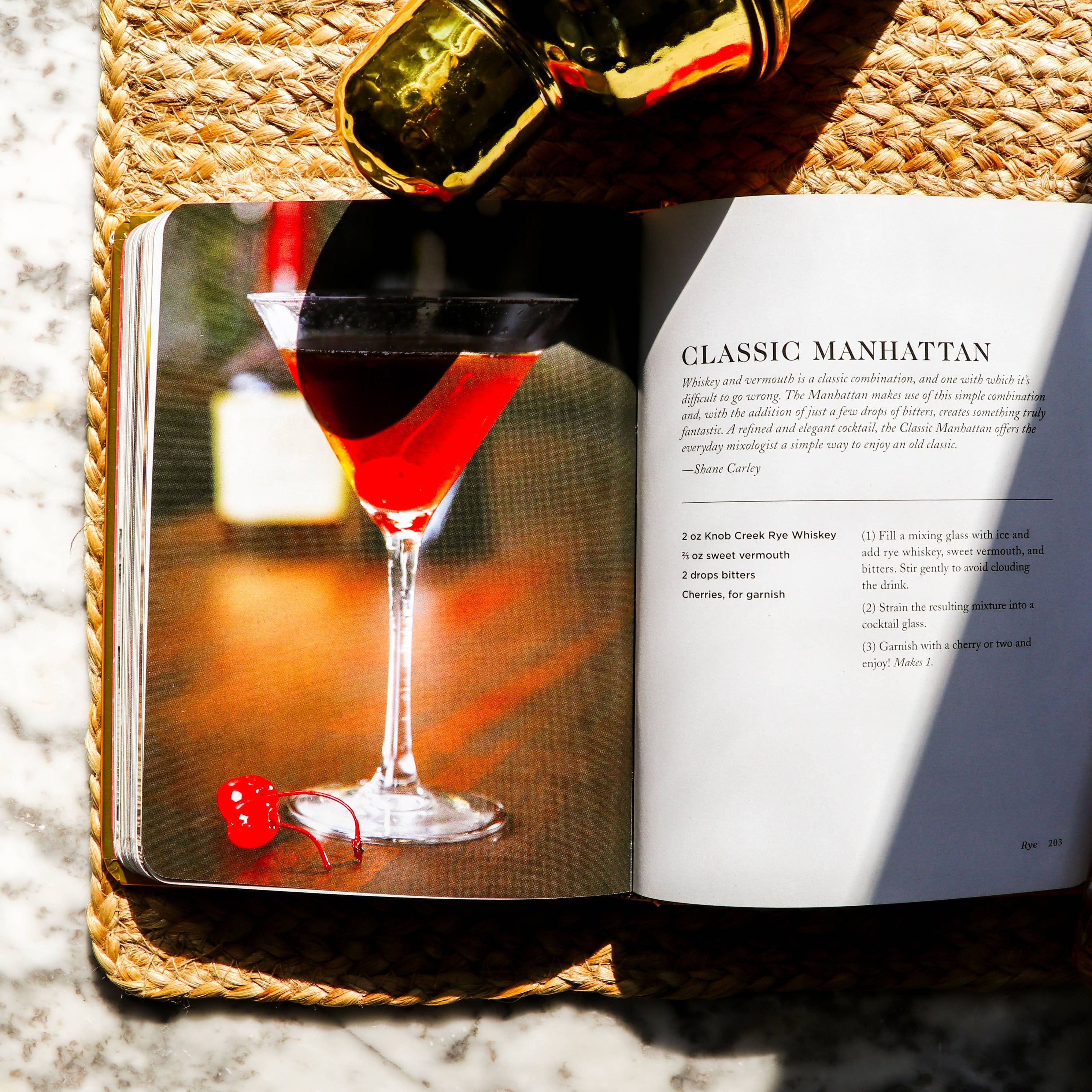Open book with a cocktail recipe, bottle of bourbon, and glass on a woven mat.