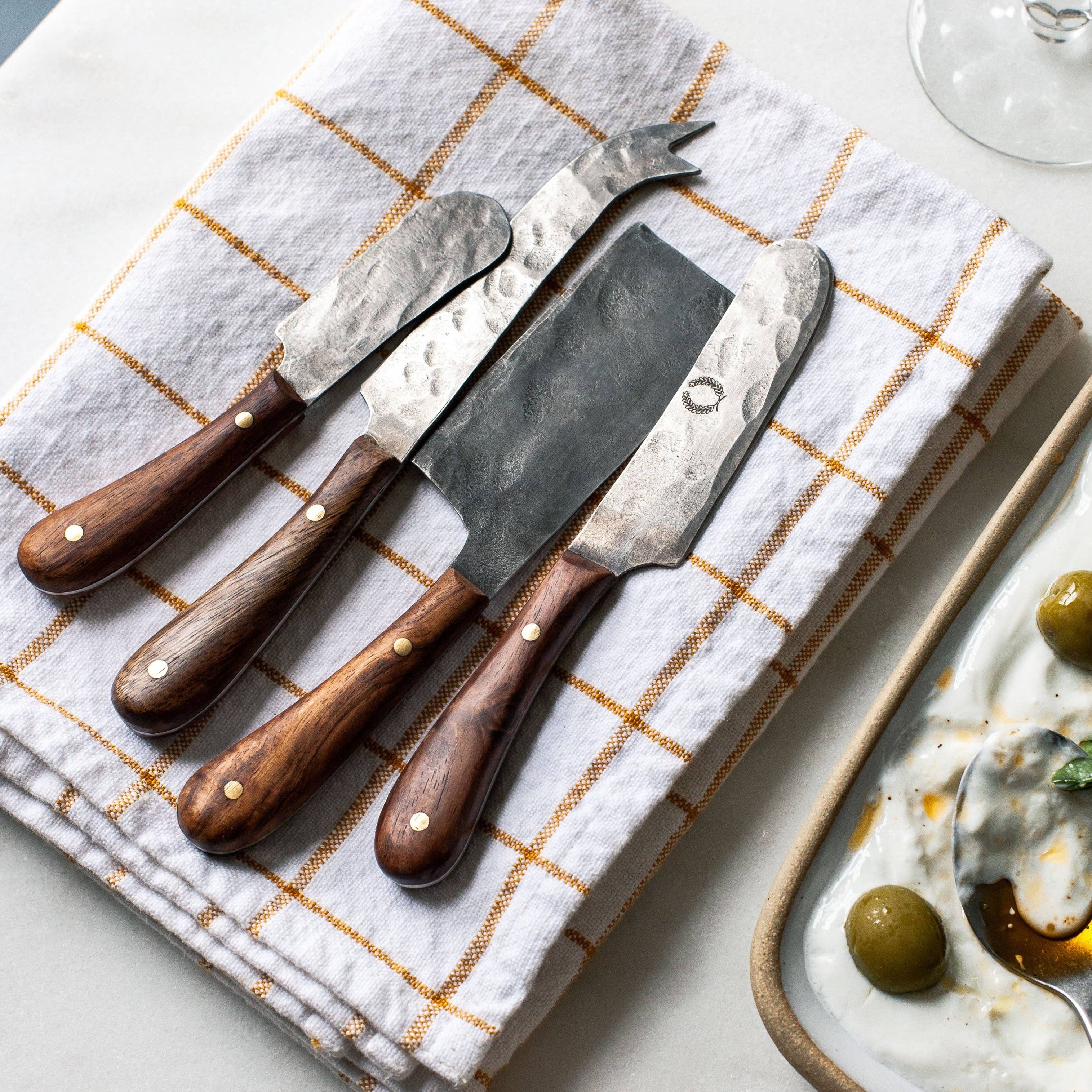 Set of three cheese knives on a checkered cloth with a plate of food and a glass of wine in the background.