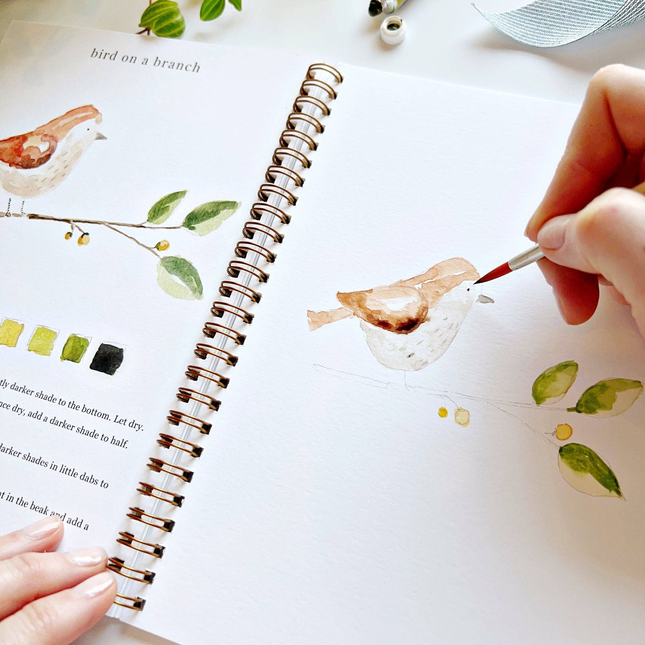 Person painting a bird illustration in a notebook with a paintbrush.