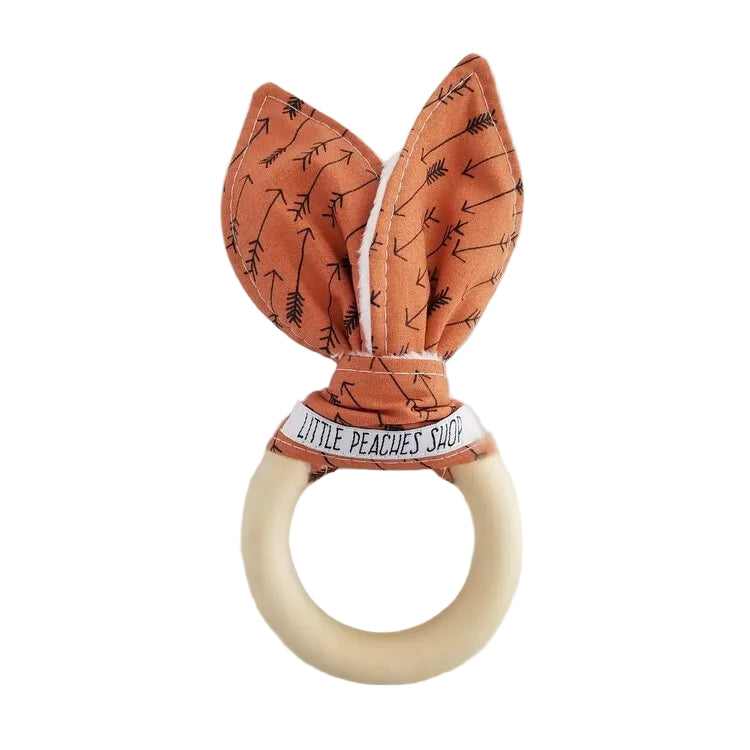 A beige-colored baby teether with an orange fabric attached to a wooden ring, decorated with clay arrows patterns.